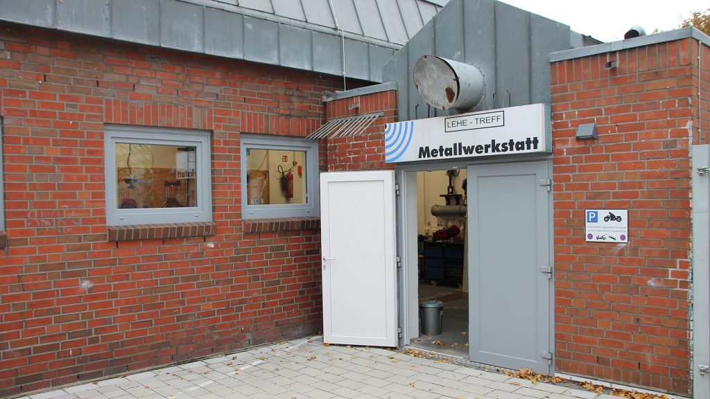Ein Gebäudeeingang von Außen. Über der Tür hängt ein großes Schild auf dem steht "Metallwerkstatt".
