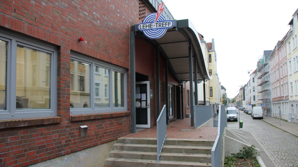 Ein Gebäude von Außen. Ein Treppenaufgang führt zur Eingangstür. Über dem Eingang hängt ein Reklame-Logo. Das Logo besteht aus einer blauen Zielscheibe mit einem roten Pfeil.