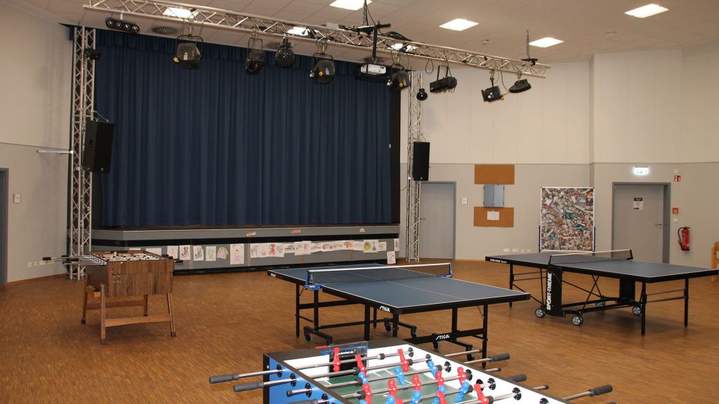 Ein großer Raum mit Spielgeräten wie Tischtennisplatten und Tischkicker. Zental im Bild ist eine Bühne mit Scheinwerfern zu sehen.