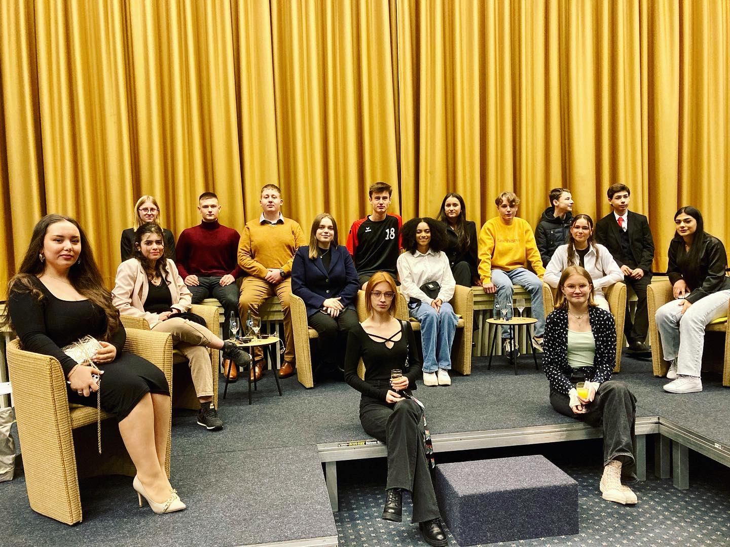 In dem Bild sieht man 16 Jugendliche, die Mitglieder des Jugendparlaments sind. Sie sitzen auf einer Bühne, am Bühnenrand oder auf Stühlen auf der Bühne. Hinter Ihnen ist ein gelber Vorhang zu sehen.